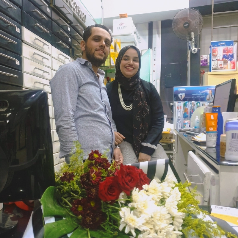 Dr. Noor Abdelbasst and her husband Mostafa standing behind the counter of their first pharmacy branch next to a fresh bouquet of flowers.