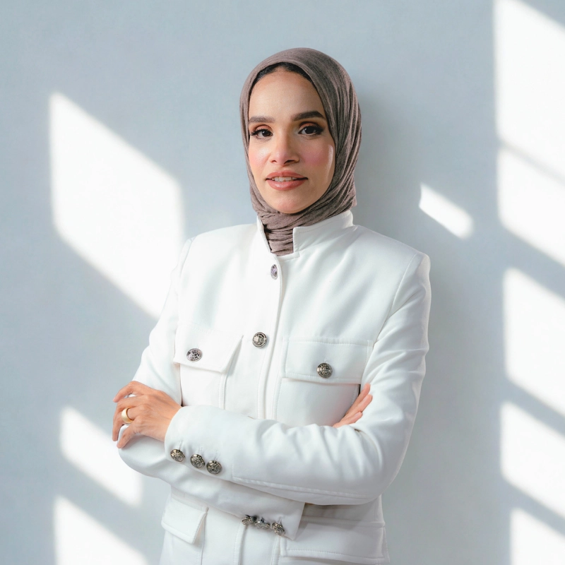 A professional studio portrait of Dr. Noor Abdelbasst smiling with crossed arms, wearing a clean white blazer and a mauve hijab.