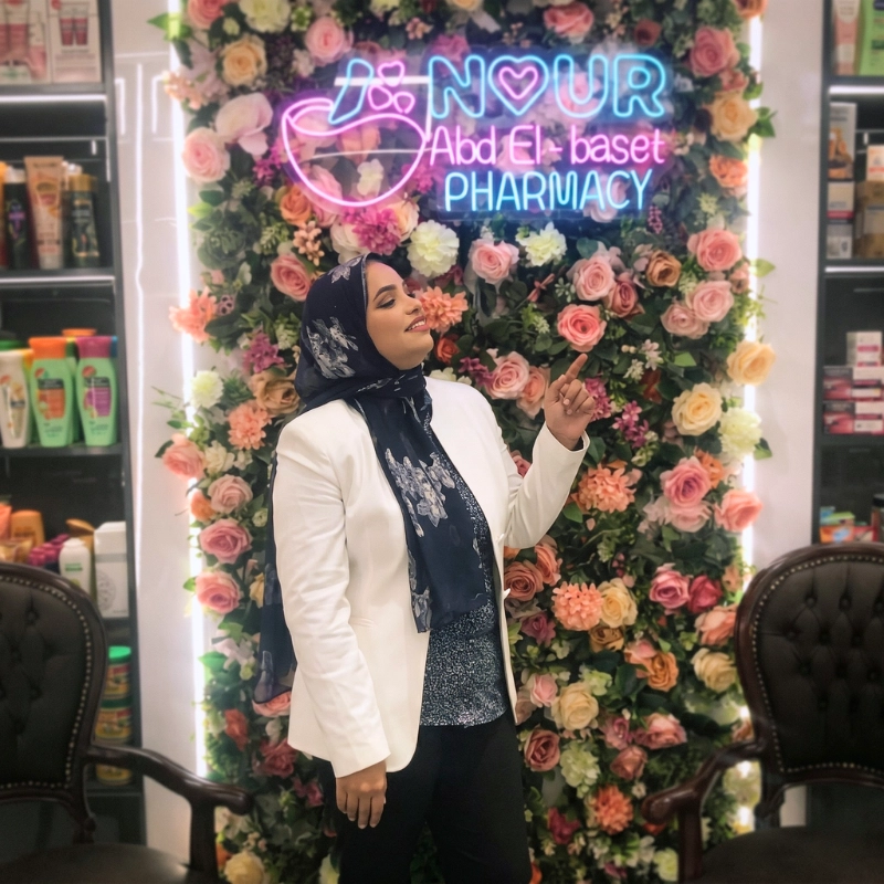 Dr. Noor Abdelbasst smiling and pointing at a glowing pink and blue neon sign that reads Nour Abd El-baset Pharmacy, backed by a wall full of colorful roses.