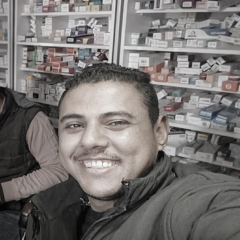 A close-up smiling portrait of Mosaab, the first delivery captain of Noor Abdelbasst Pharmacy, with shelves of medicine in the background.