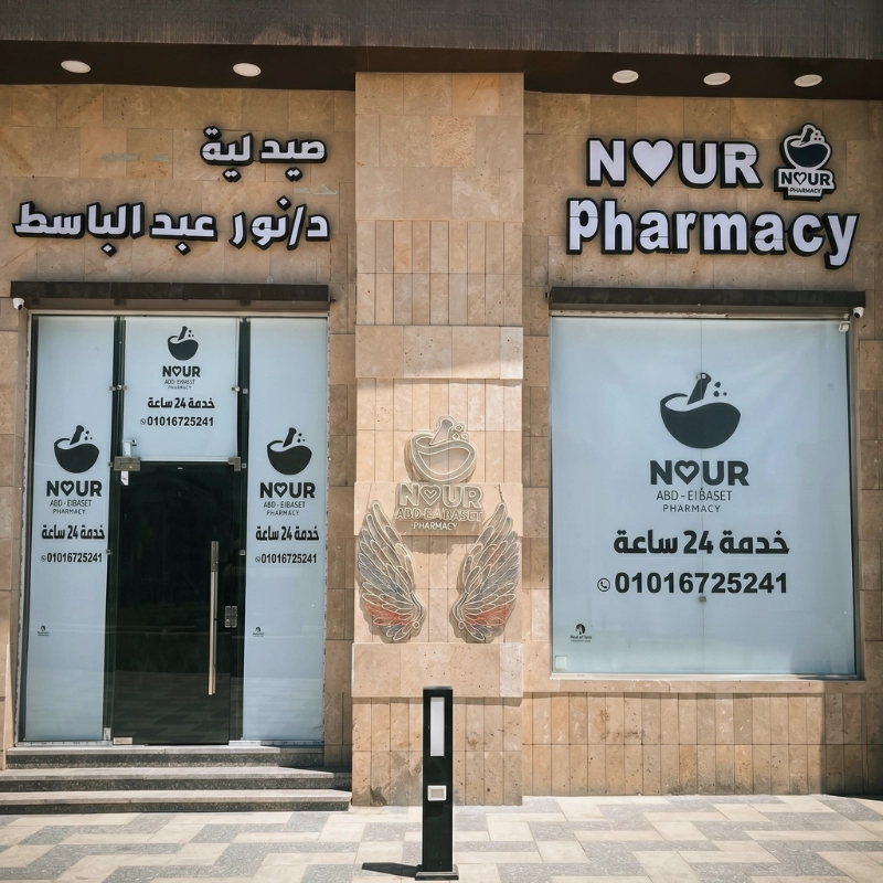 The modern exterior stone facade of Noor Abdelbasst Pharmacy showing the 24-hour service signs and the gold wings brand logo.
