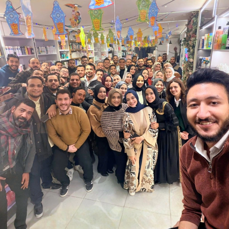 A large group selfie of the smiling Noor Abdelbasst Pharmacy team standing together inside a branch decorated with colorful Ramadan lanterns.
